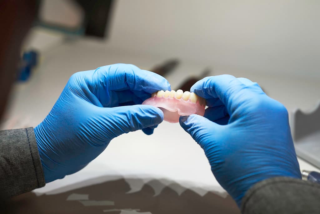 A technician's gloved hands holding a finished digital denture base with set teeth under laboratory light.